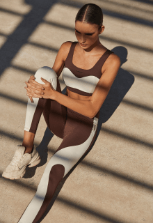 mulher se alongando entre brechas de luz e sombras, ela está usando uma roupa de ginástica com calça e top nas cores bege e branco